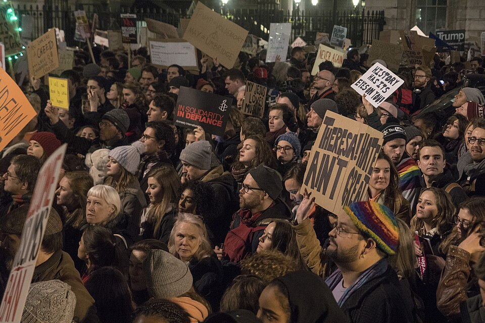 A large crowd of people participates in a 2017 protest, holding up various signs with messages like IN SOLIDARITY IN DEFIANCE and REFUGEES WELCOME. The crowd is diverse and gathered closely together at night.
