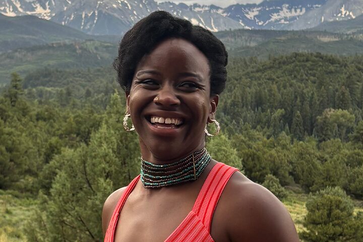 A woman wearing a coral sleeveless top, beaded necklace, and hoop earrings smiles outdoors, with a lush green forest and snow-capped mountains in the background.