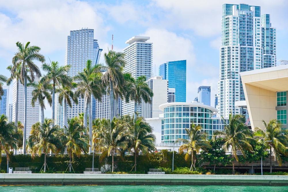 A waterfront view of downtown Miami with tall modern skyscrapers, numerous palm trees, and turquoise water in the foreground under a partly cloudy sky.