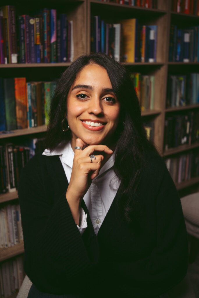 A woman with long dark hair smiles and rests her chin on her hand, sitting in front of bookshelves filled with colorful books. She wears a black sweater over a white collared shirt.