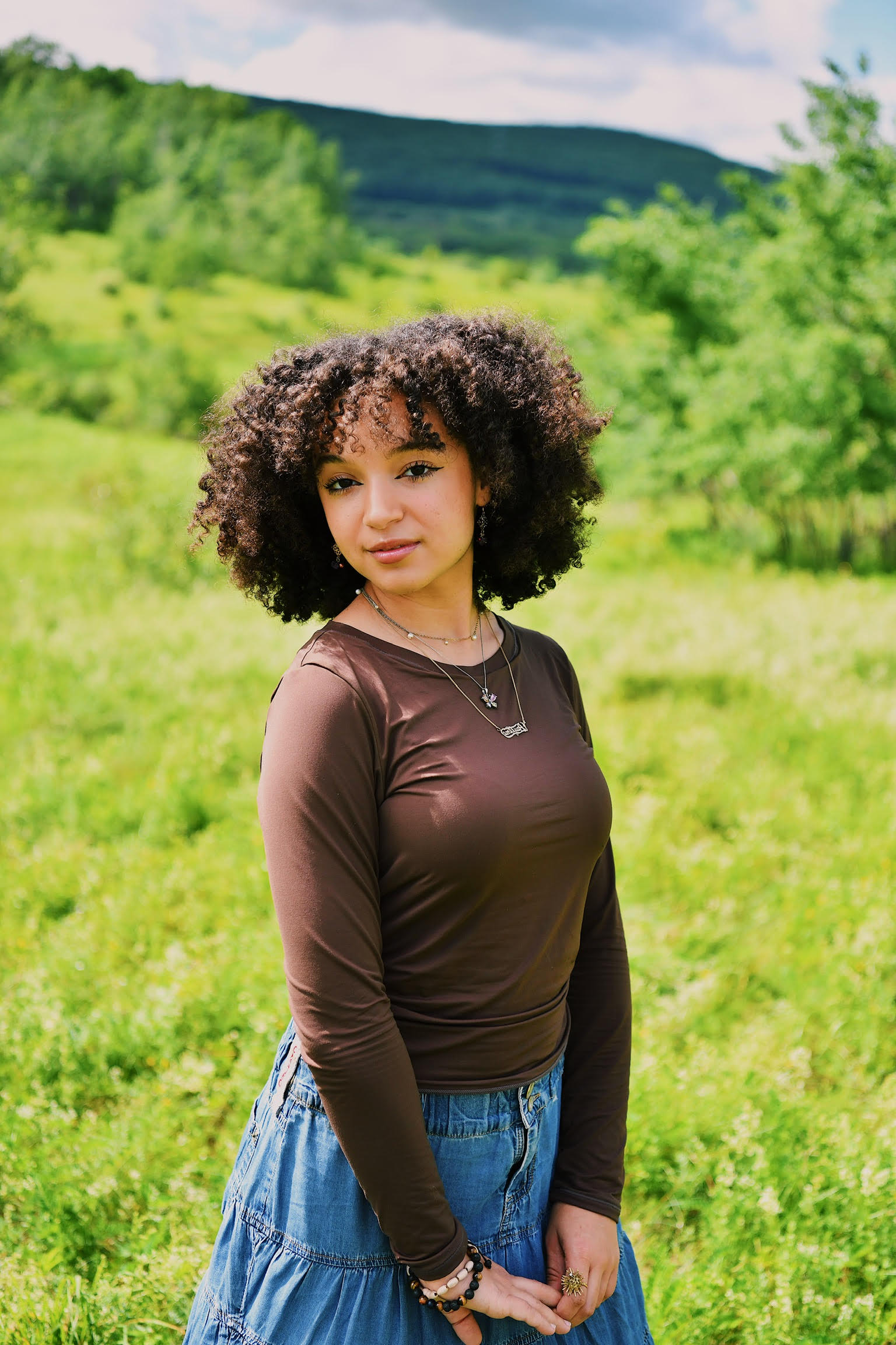 A young woman with curly hair, wearing a brown long-sleeve shirt and a blue skirt, stands in a green field with trees and hills in the background on a sunny day.