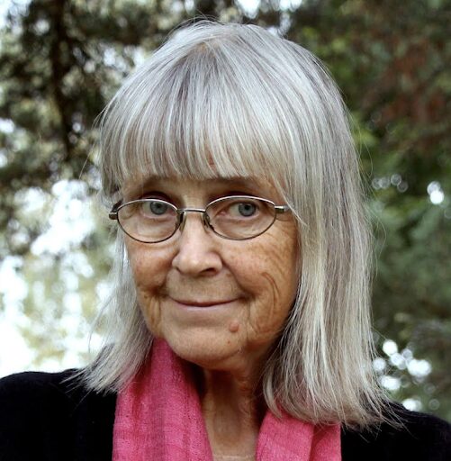 An older woman with gray hair and glasses, wearing a pink scarf and a dark top, stands outdoors in front of blurred trees, smiling gently at the camera.