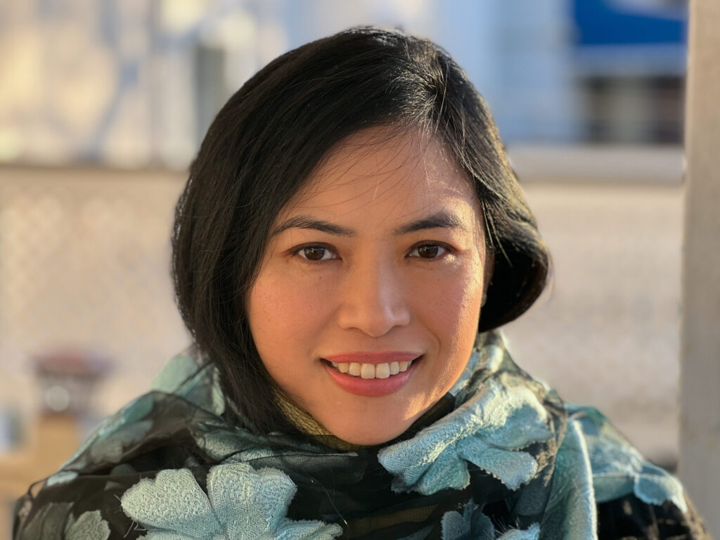 A woman with black hair smiles at the camera, wearing a dark floral-patterned scarf. The background is softly blurred with warm sunlight illuminating her face.
