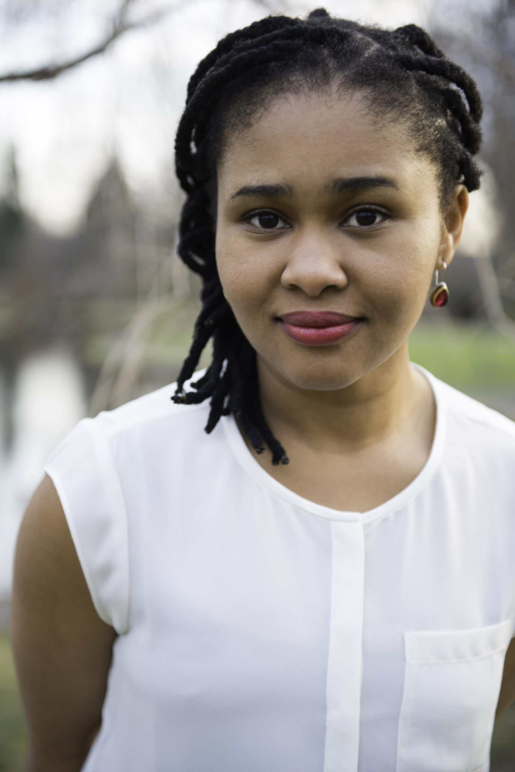 A woman with black dreadlocks, wearing a white sleeveless blouse and red earrings, stands outdoors with a soft smile. The background is blurred, showing greenery and water.