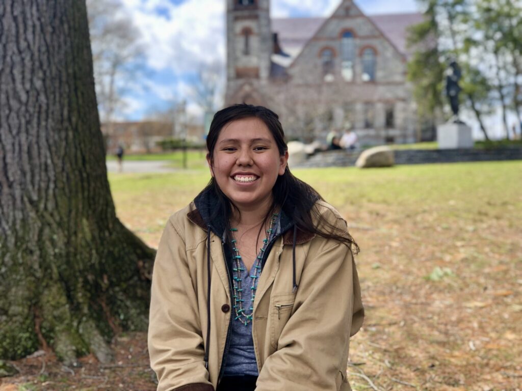 A young woman sits and smiles in a tan jacket and turquoise necklace near a large tree, with a historic stone building and green lawn in the background on a bright, partly cloudy day.