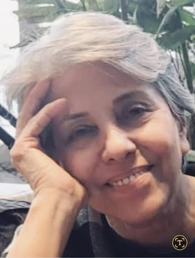 An older woman with short gray hair smiles gently, resting her head on her hand. Green plants are visible in the blurred background.