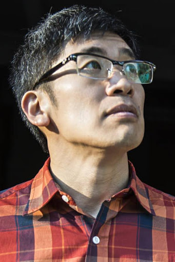 A man wearing glasses and a red plaid shirt looks upwards with a serious expression, against a dark background.