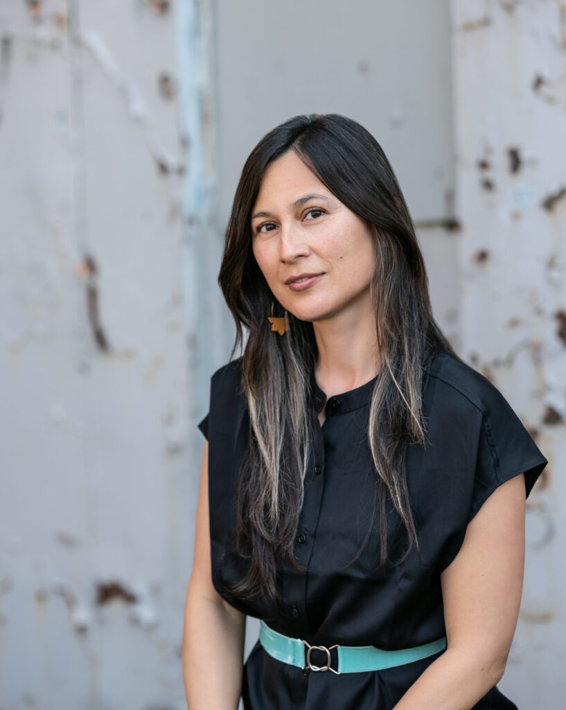 A woman with long dark hair and streaks of gray, wearing a black button-up dress with a light blue belt, stands in front of a weathered, light-colored metal wall. She gazes gently toward the camera.