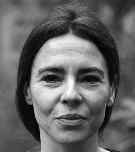 Black and white close-up portrait of a woman with straight dark hair, parted in the center, looking directly at the camera with a neutral expression. The background is blurred.
