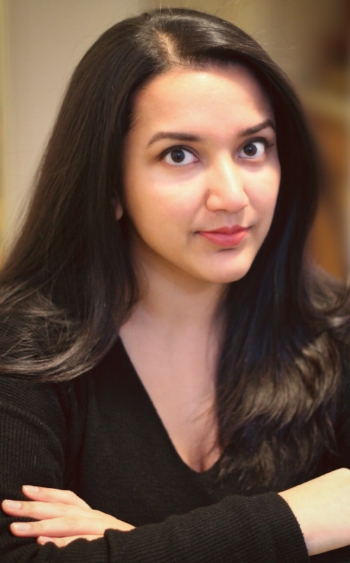 A woman with long, dark hair and a black top sits with her arms crossed, looking directly at the camera with a neutral expression. The background is softly blurred.