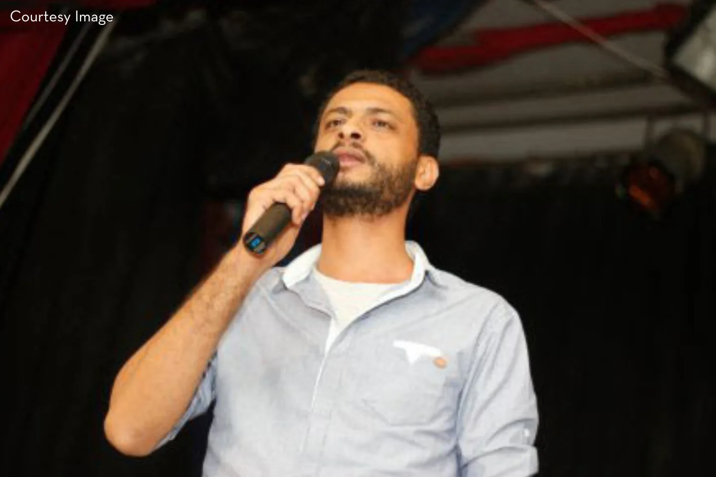 A man with short curly hair and a beard holds a microphone and speaks onstage. He wears a light blue button-up shirt. The background is dark with some indistinct structures and red elements.