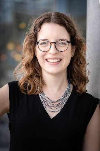 A smiling young woman with wavy brown hair and glasses, wearing a black top and a layered silver necklace, stands in front of a blurred background.