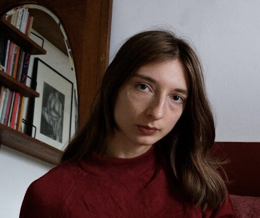 A young woman with long brown hair and a serious expression wears a red sweater, sitting indoors near a bookshelf and a framed portrait.