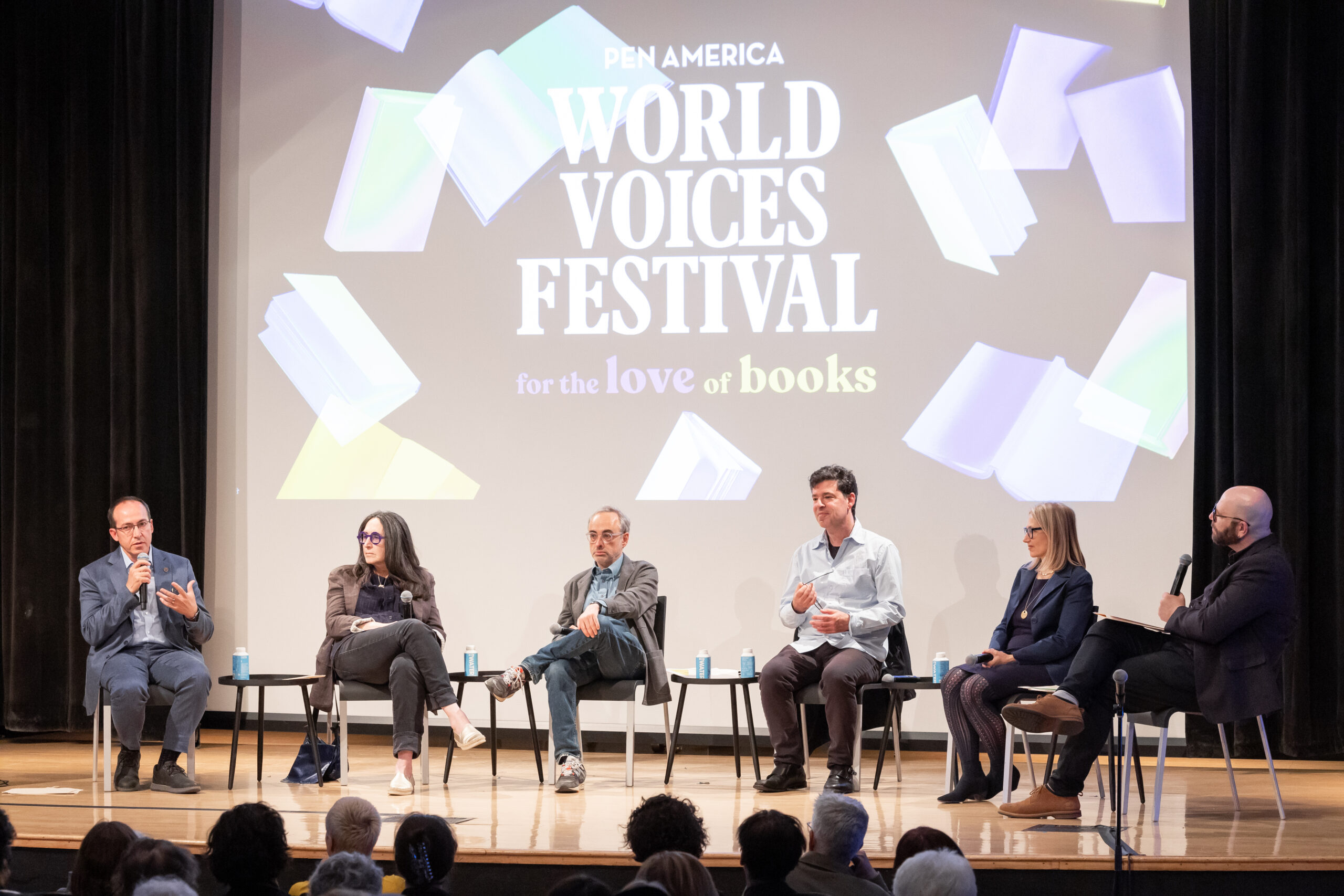 Six people sit on stage in front of a screen that reads “PEN America World Voices Festival for the love of books,” participating in a panel discussion while an audience watches.