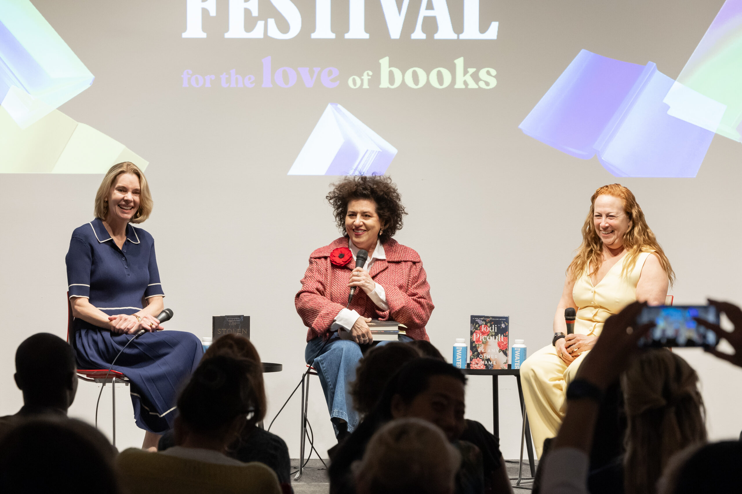 Three women sit on stage in front of an audience at a book festival, smiling and speaking. Two books and water bottles are on the table between them, with a festival logo projected behind. An audience member holds up a phone.