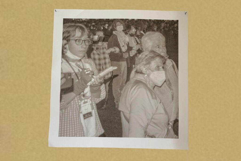 A black-and-white photo shows a group of people, some wearing masks and scarves, standing closely together. One person in the foreground holds a notebook and pen, appearing to take notes. The photo is pinned to a beige wall.