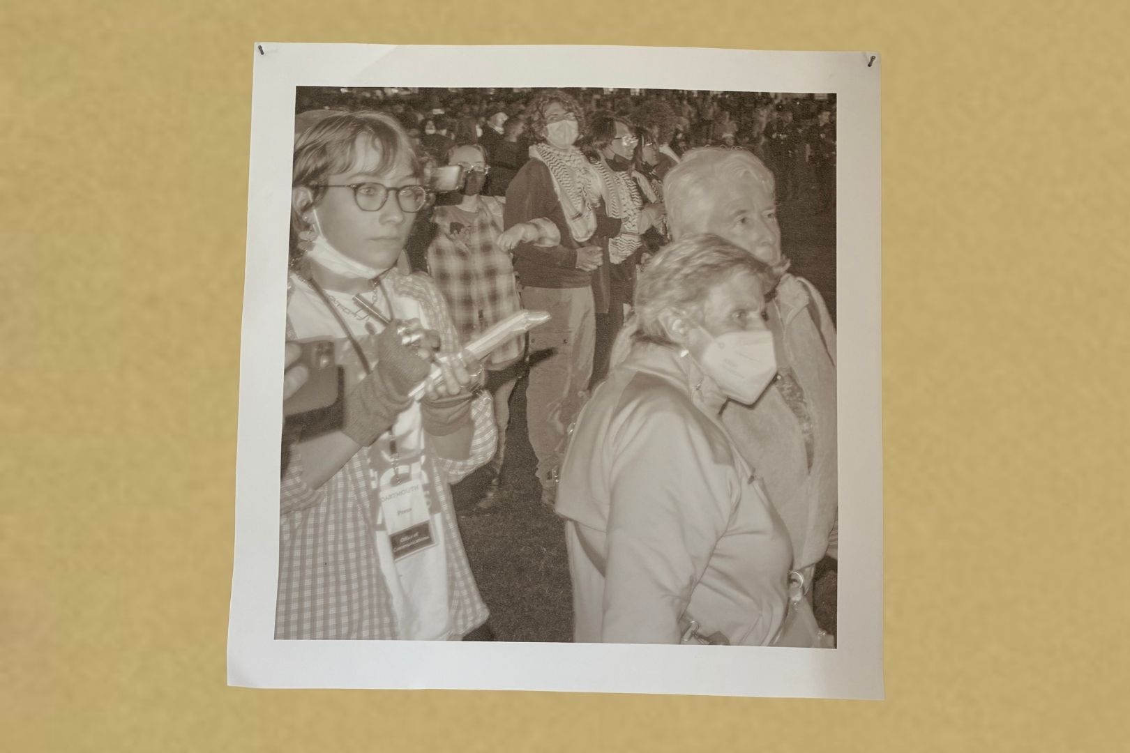 A black-and-white photo shows a group of people, some wearing masks and scarves, standing closely together. One person in the foreground holds a notebook and pen, appearing to take notes. The photo is pinned to a beige wall.