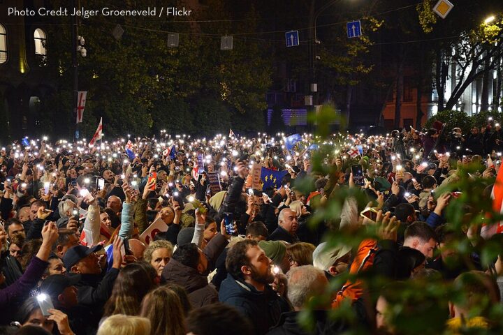 A large crowd gathers outdoors at night, many people holding up smartphones with flashlights on, creating a sea of lights. Some are waving flags, and trees are visible in the background.