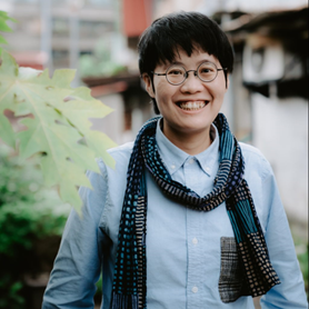 A person with short dark hair, glasses, and a blue patterned scarf smiles outdoors, wearing a light blue shirt with a checkered pocket. Green foliage and blurred buildings appear in the background.