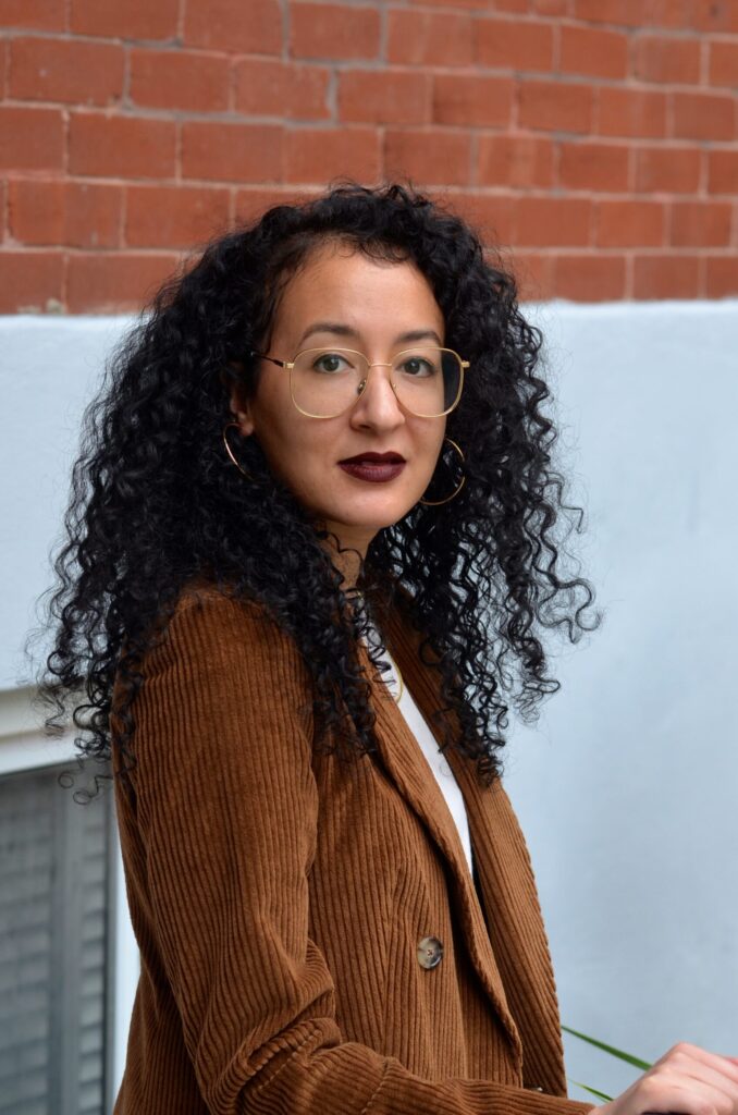 A person with long, curly black hair, wearing gold-rimmed glasses, hoop earrings, and a brown corduroy blazer, stands in front of a brick and white wall, looking confidently at the camera.