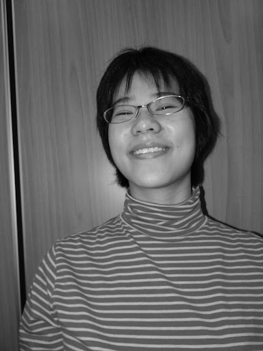 A person with short hair and glasses smiles while wearing a striped turtleneck shirt, standing in front of a wooden panel background. The image is in black and white.