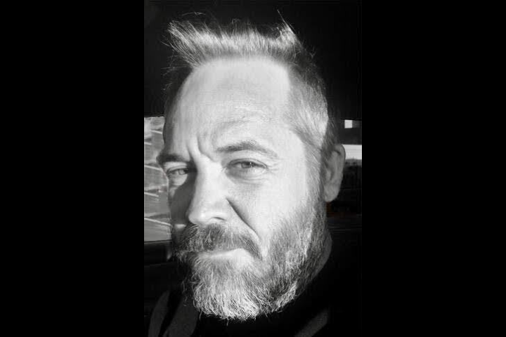 Black and white close-up of a bearded man with short hair, looking toward the camera with a serious expression. Strong sunlight casts shadows across his face.