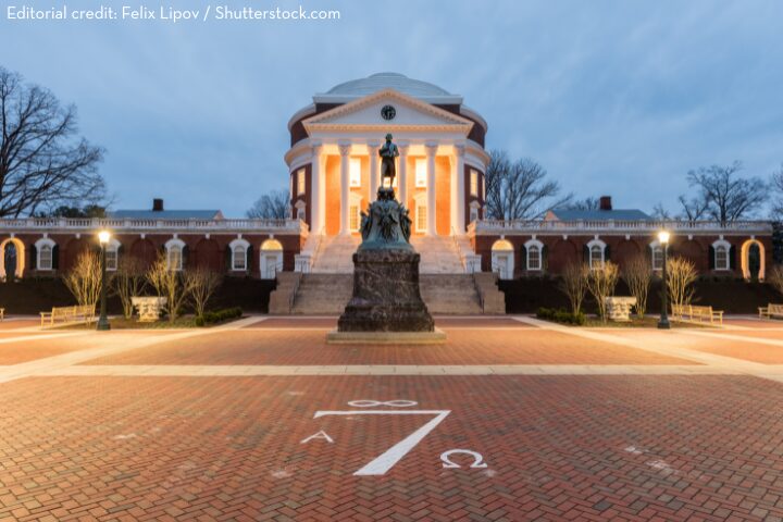 The Rotunda at the University of Virginia is lit up at dusk, with a statue in front and Greek symbols painted on the brick plaza. Leafless trees and benches line the walkway.