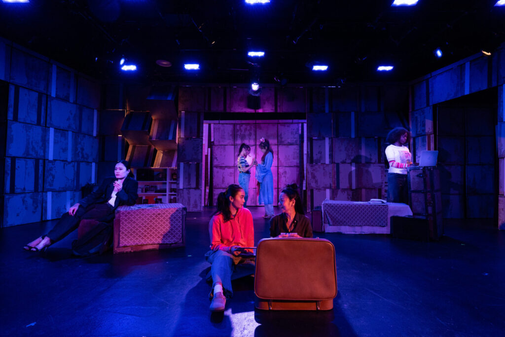 Five people are on stage, illuminated by blue and purple lighting. Two sit on suitcases at the front, talking. Three others are scattered in the background, with one standing at a table with a laptop. The set features a geometric backdrop.