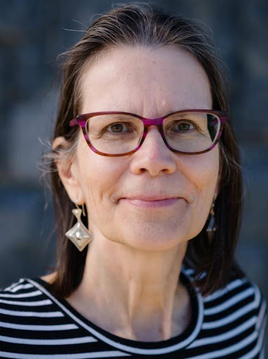 A middle-aged woman with long brown hair, wearing glasses with purple frames, dangling geometric earrings, and a black-and-white striped top, smiling softly at the camera with a blurred outdoor background.