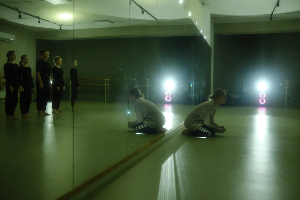 Five people in dark outfits stand by a mirrored wall in a dance studio. One person sits on the floor near the mirror, facing the wall, with bright lights shining in the background, reflecting off the floor and glass.
