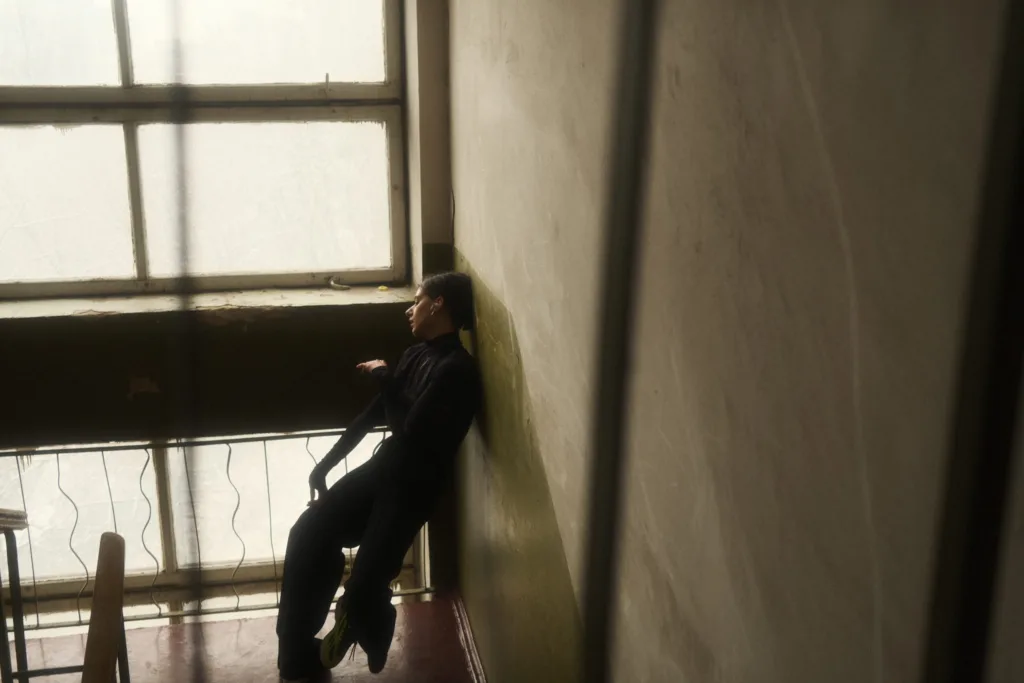 A person in dark clothing leans against a wall in a dimly lit room, next to a large window with soft light coming through, creating a moody and contemplative atmosphere.