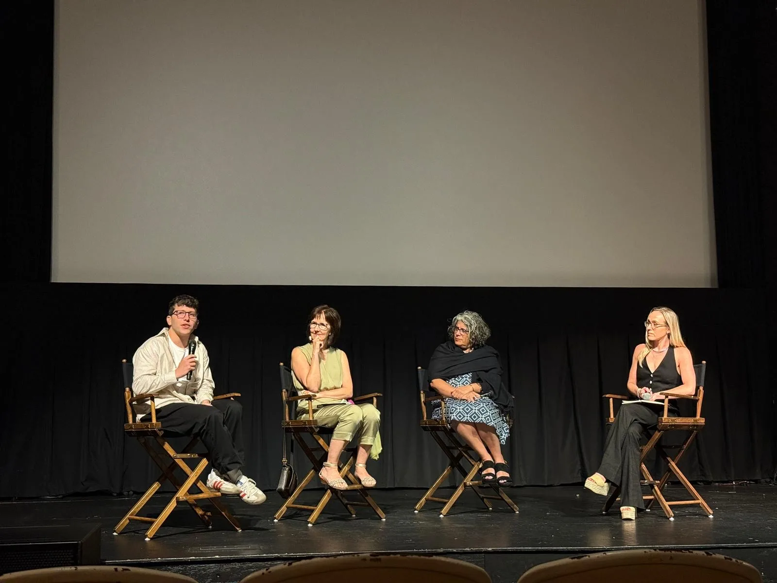 Four people are seated on stage in director’s chairs, having a panel discussion in front of a large blank screen. One person speaks into a microphone while the others listen attentively. The setting appears to be a theater or cinema.