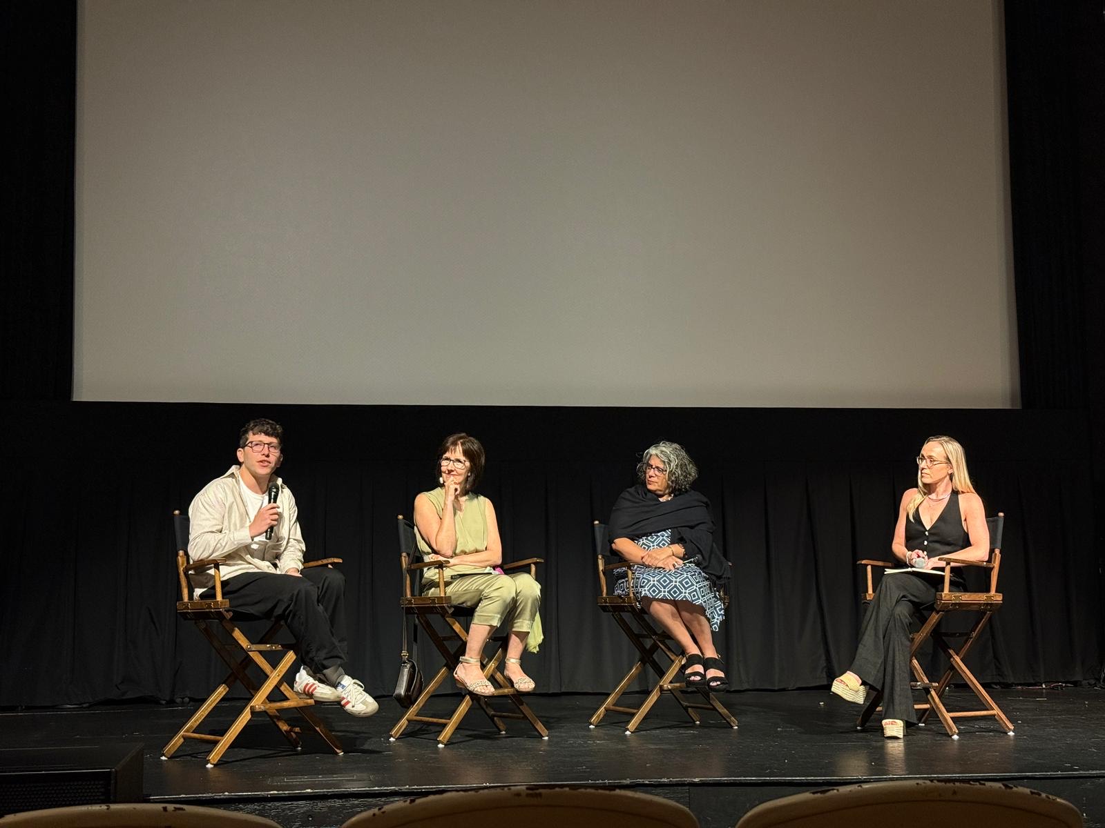 Four people are seated on stage in director’s chairs, having a panel discussion in front of a large blank screen. One person speaks into a microphone while the others listen attentively. The setting appears to be a theater or cinema.