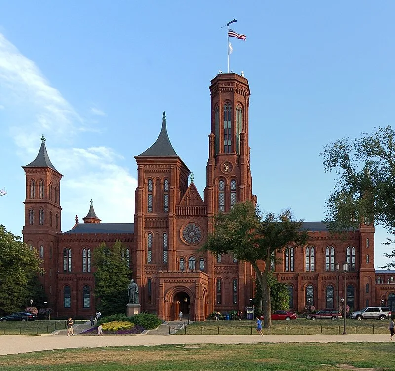 Red brick Smithsonian Institution Building, known as the Castle, with tall towers and a central rose window, stands under a blue sky; people walk and relax on the lawn in front.