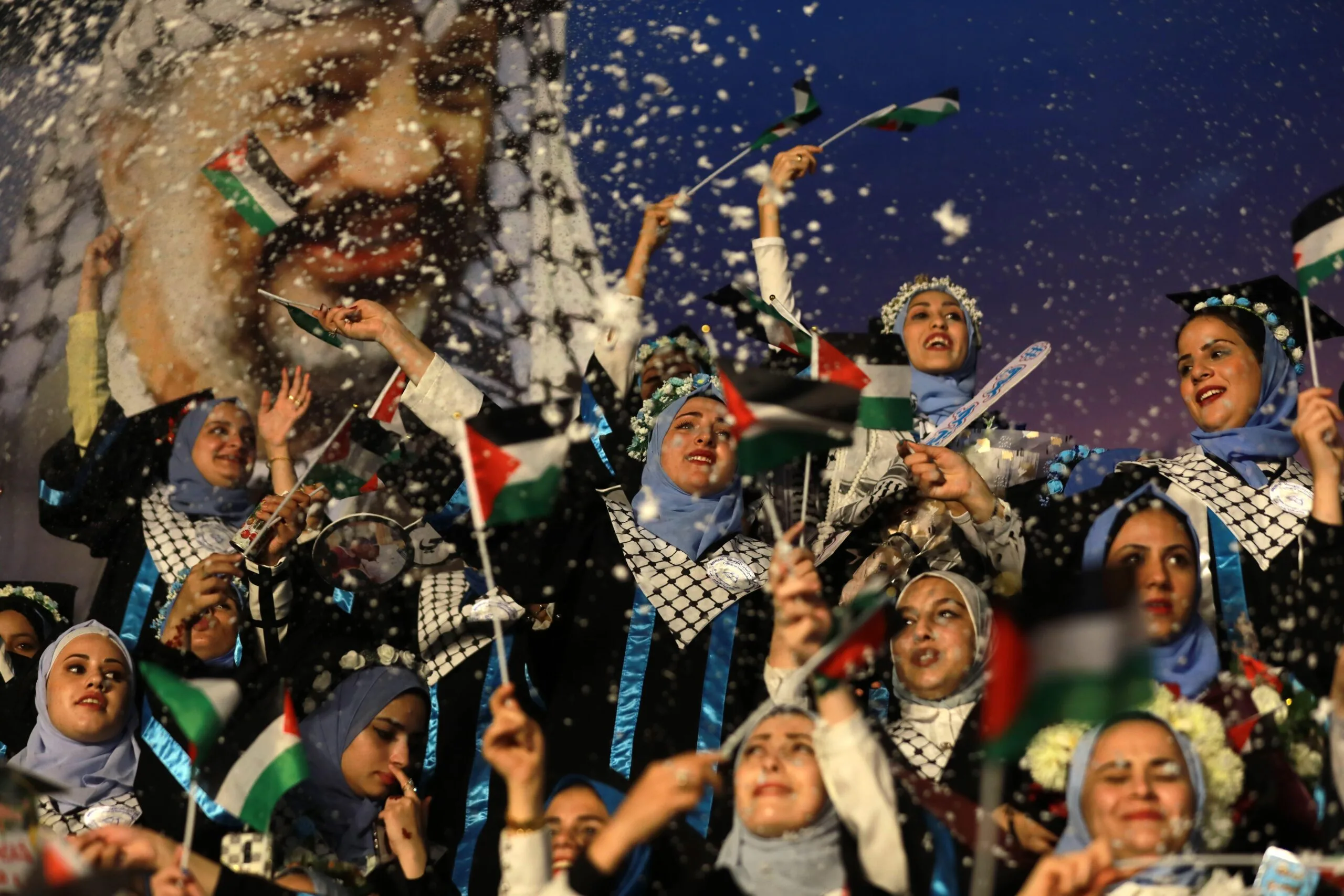 A group of women in traditional dress wave Palestinian flags and celebrate under falling confetti, with a large portrait of a smiling man in the background.