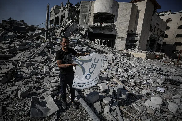 A man stands amid rubble, holding a damaged sign with OCSC text. Behind him, extensive destruction is visible, with collapsed buildings and debris scattered everywhere under a cloudy sky.