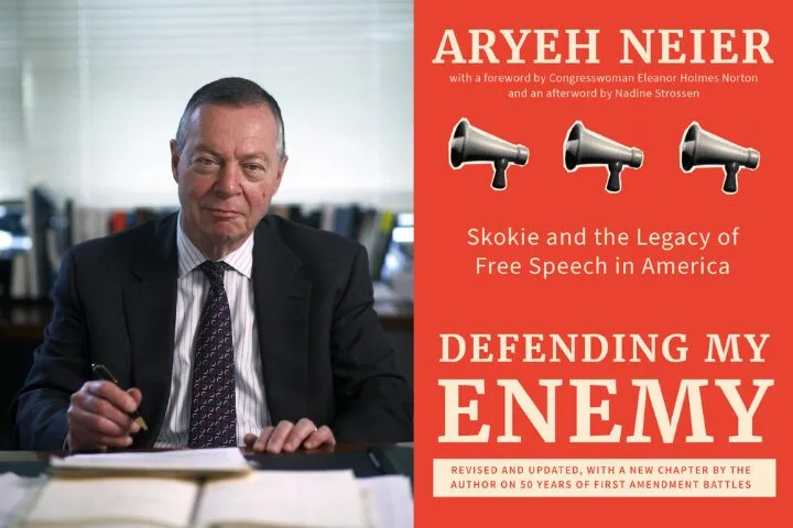 A man in a suit sits at a desk writing, with the book cover for Defending My Enemy by Aryeh Neier beside him. The cover is orange with megaphone icons and white and yellow text about free speech and Skokie.