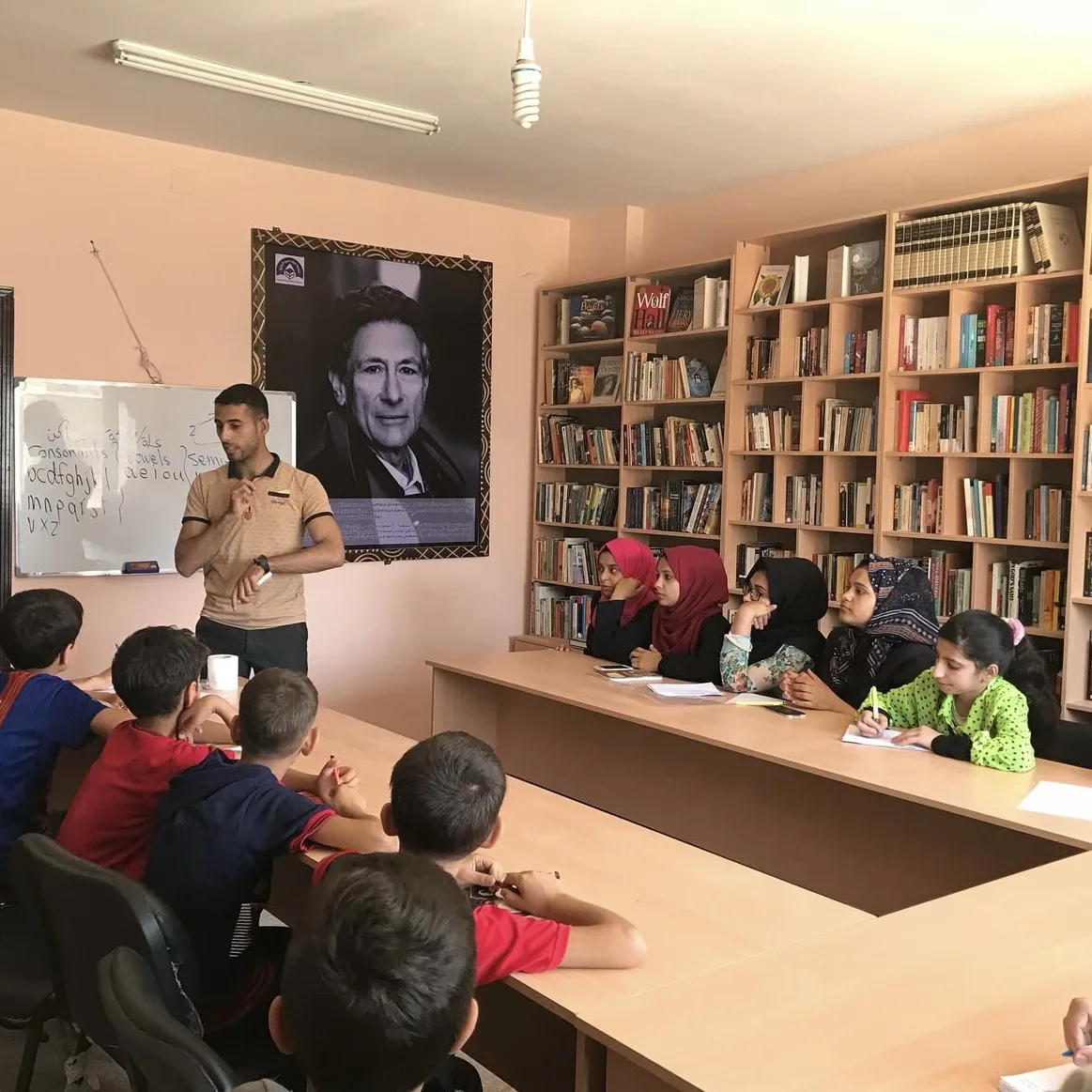 A teacher stands by a whiteboard teaching a group of children seated at tables in a library. The children are listening attentively. Bookshelves line the walls and a large portrait hangs in the background.