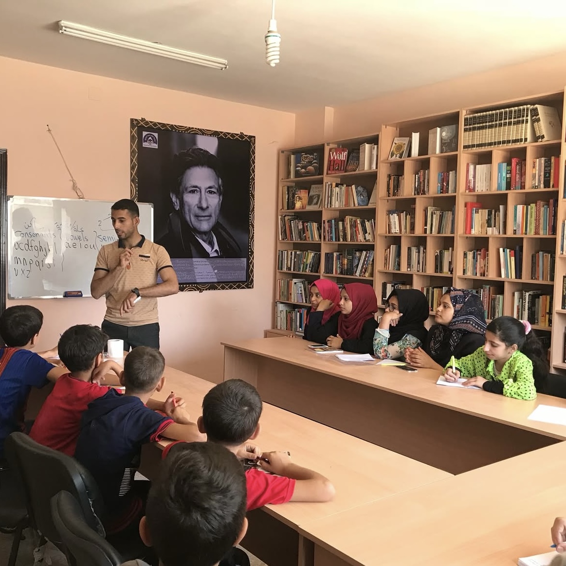 A teacher stands by a whiteboard teaching a group of children seated at tables in a library. The children are listening attentively. Bookshelves line the walls and a large portrait hangs in the background.