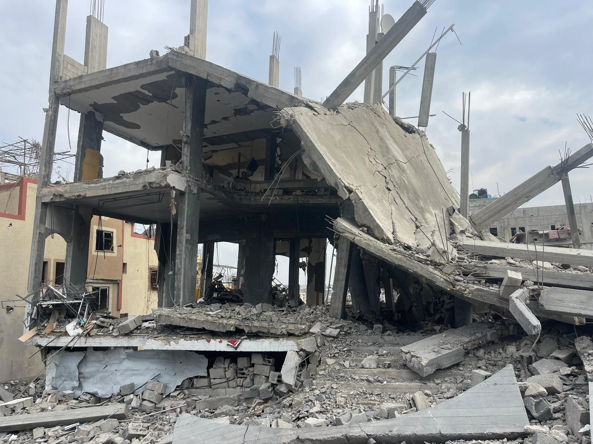 A heavily damaged concrete building with collapsed floors and exposed supports, surrounded by debris and rubble under a cloudy sky.