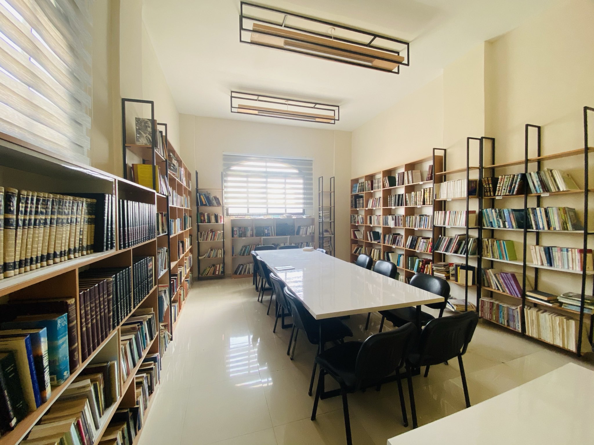 A bright library room with shelves full of books on both sides, a large white table in the center surrounded by black chairs, and sunlight streaming in through a window at the far end.