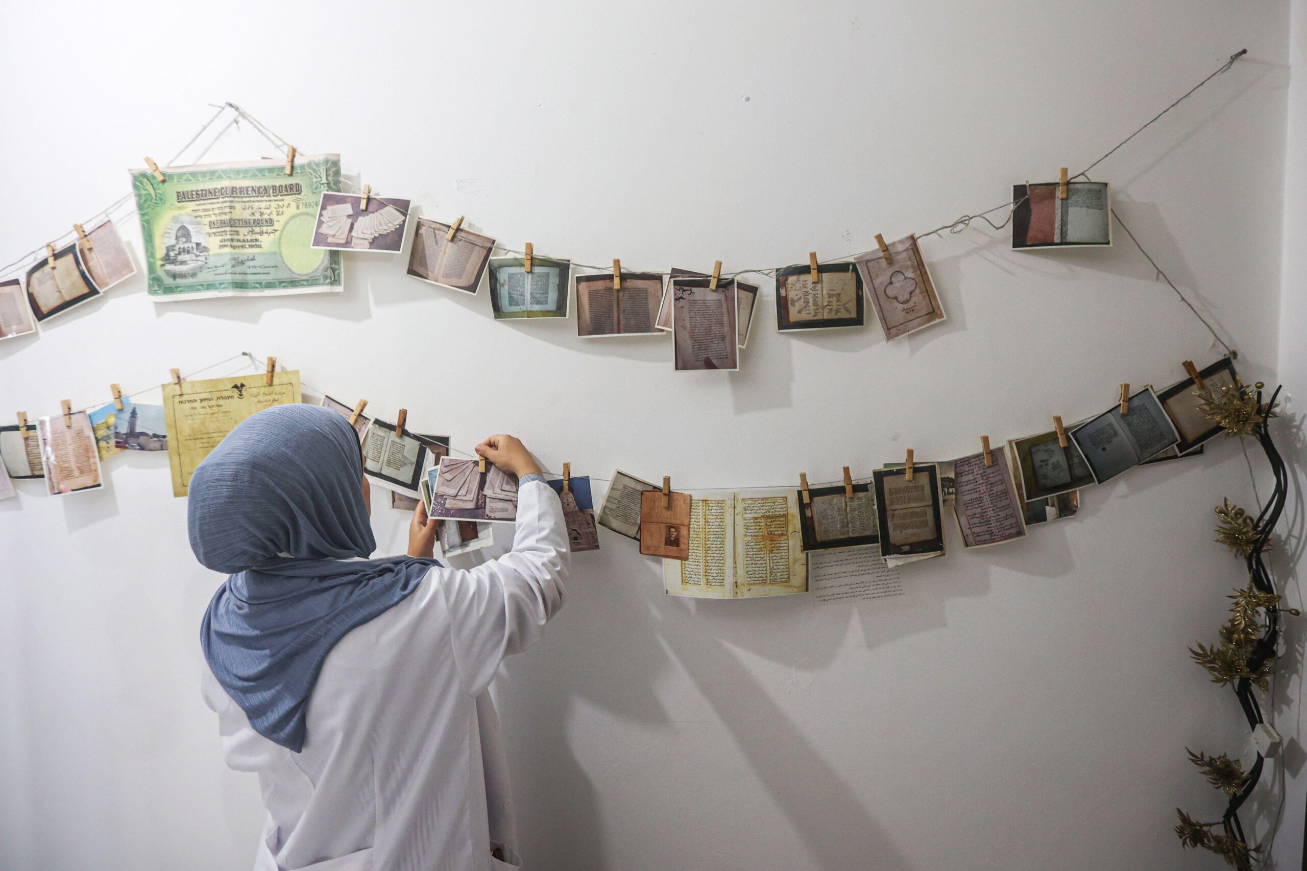 A person wearing a grey hijab and white shirt hangs or adjusts photos and documents on string with clothespins, displayed against a white wall.