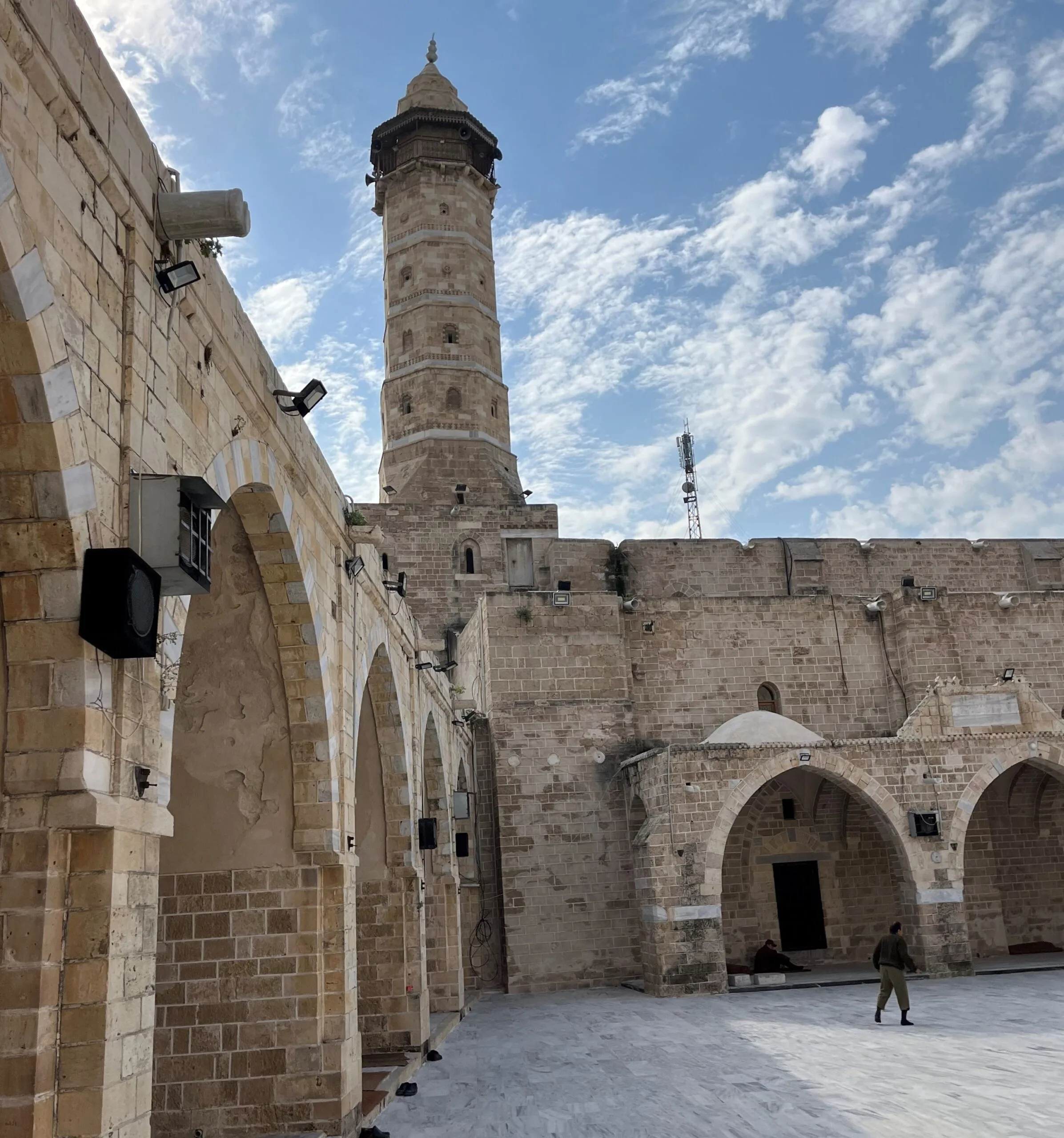 Historic stone courtyard with arched doorways and a tall, cylindrical minaret under a partly cloudy sky. A person walks across the open space, and the ancient walls show signs of age and weathering.