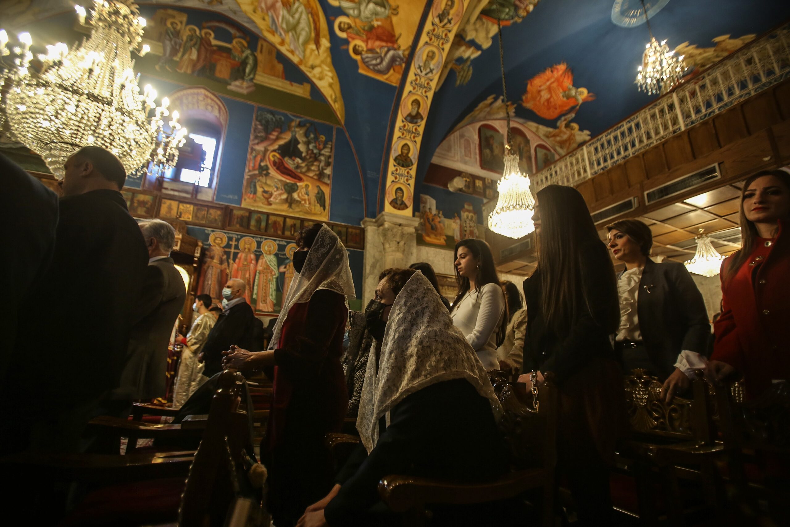 People stand and sit in a richly decorated church with ornate chandeliers and colorful religious murals. Some women wear white lace veils. The atmosphere is solemn and reverent.