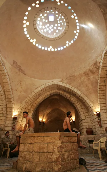 Shirtless men sit on a stone platform under a large domed ceiling with circular skylights, inside a historic, arched bathhouse with beige stone walls.