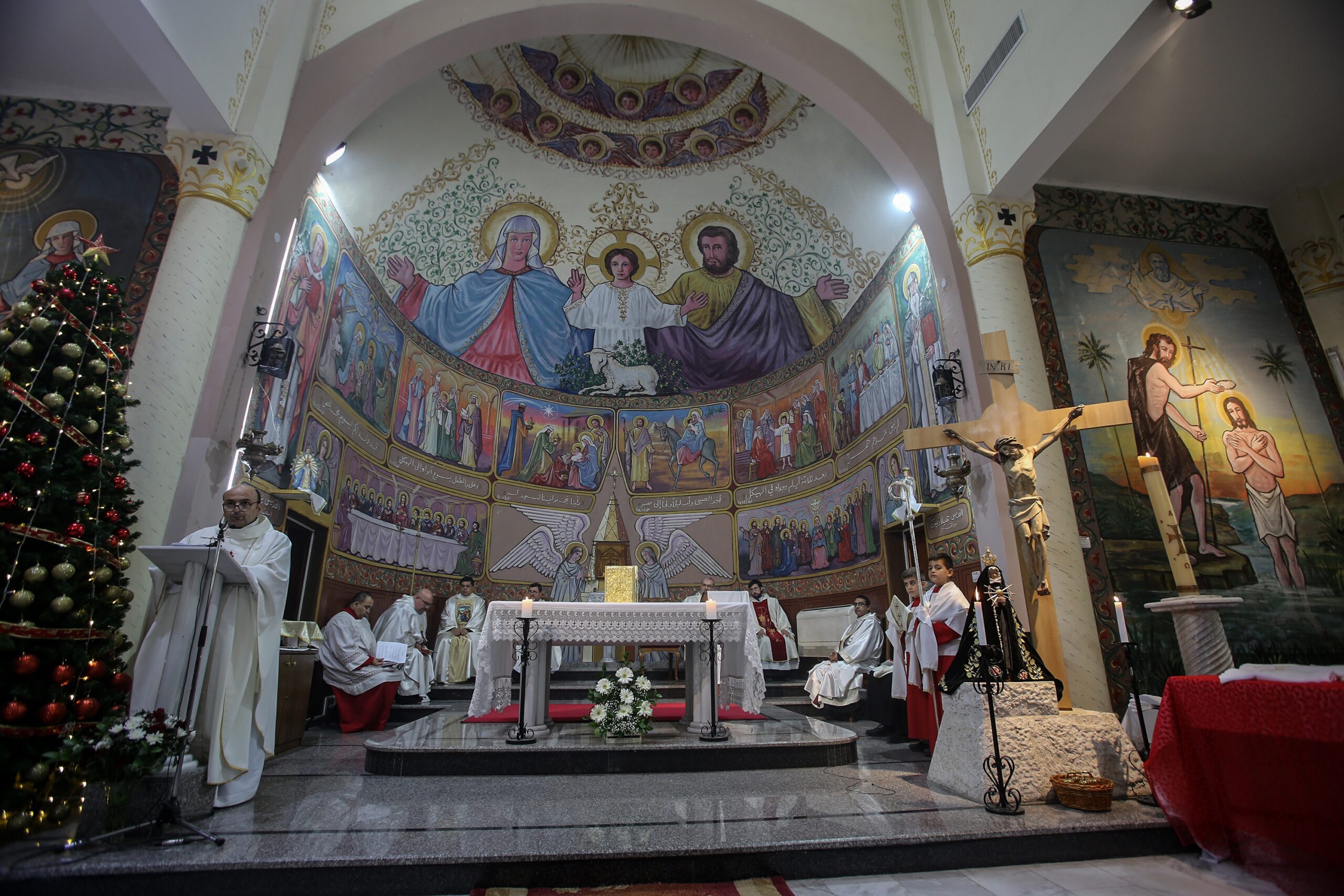 A group of priests and altar servers conduct a service in a richly decorated church, featuring a large religious mural behind the altar and a Christmas tree to the left.