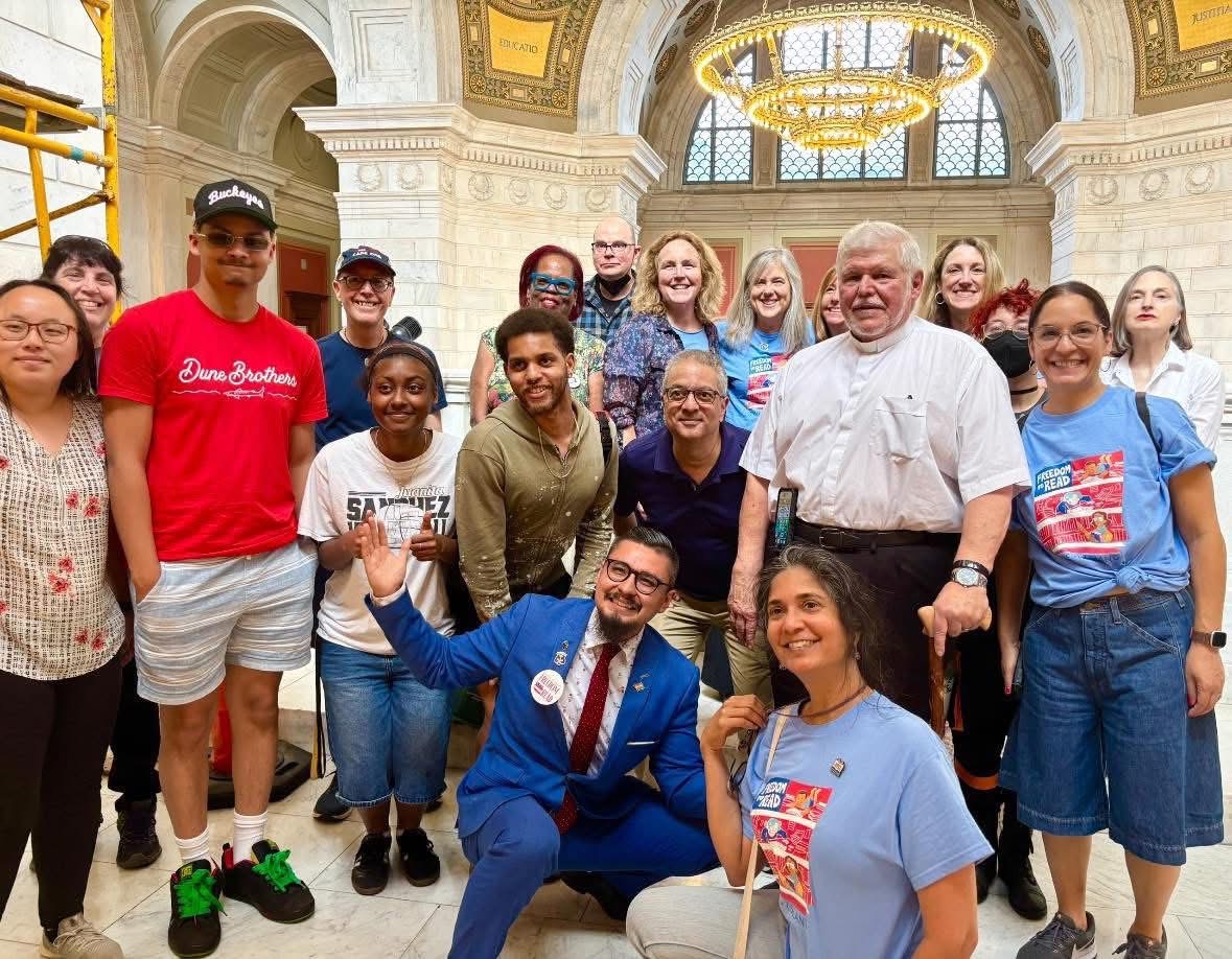 A diverse group of people, including men and women of various ages and backgrounds, smile and pose together inside an ornate hall with arched windows and a large chandelier overhead.