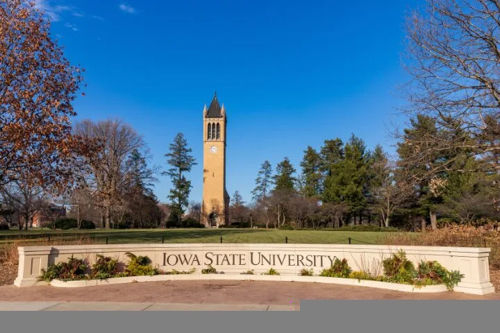The image shows the Iowa State University entrance sign with the iconic campanile clock tower in the background, surrounded by trees and clear blue sky.