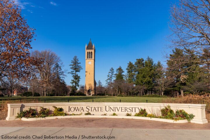 The image shows the Iowa State University entrance sign with the iconic campanile clock tower in the background, surrounded by trees and clear blue sky.