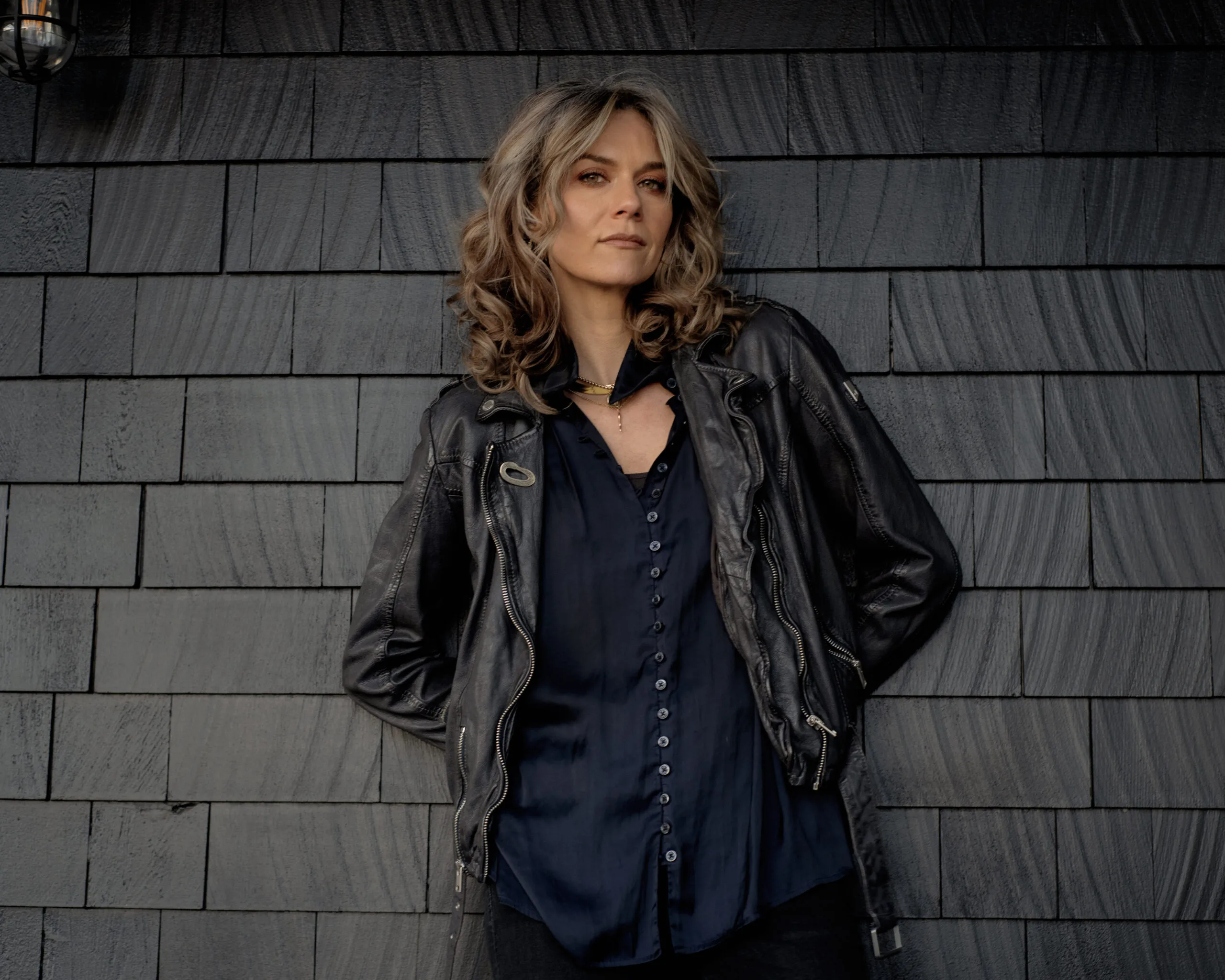 A woman with wavy, shoulder-length hair stands against a dark shingle wall, wearing a black leather jacket over a navy blue button-up shirt, looking confidently at the camera.